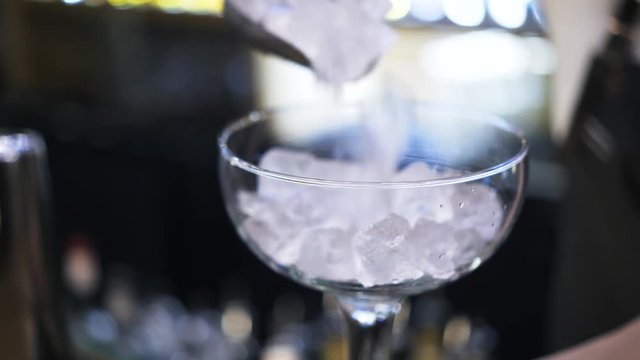 Close up of an unrecognizable and young woman bartender putting ice cubes into a cocktail glass and placing it on the counter. Handheld real time close up shot