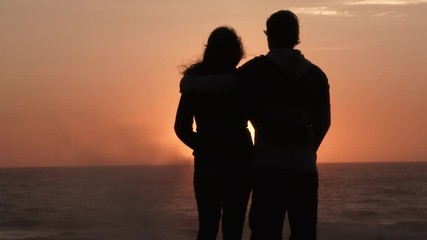 A couple in love watch the sunset and kiss at the Pacific Coast, slow motion silhouette.