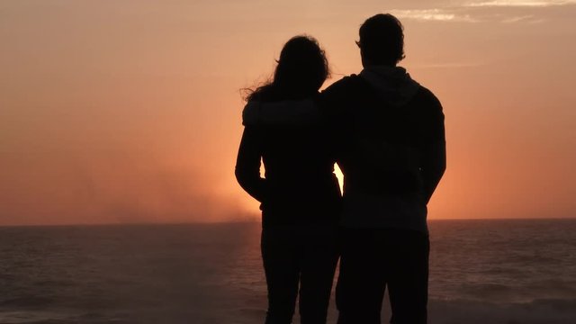 A couple in love watch the sunset and kiss at the Pacific Coast, slow motion silhouette.