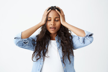 Fototapeta premium Afro-American female student with long wavy hair casually dressed feeling stressed, keeping hands on her head, with closed eyes in frustration and despair after she made serious mistake.