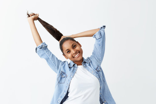 Indoor Shot Of Beautiful Afro-american Girl With Long Black Hair Gathered In Ponytail Dressed Casually Posing Indoors, Adjusting Her Hairstyle Before Going Out. People And Emotions Concept.
