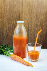 A bottle of carrot juice and a glass of carrot juice with a straw on white wood table against brown wood wall background. Still life of a healthy eating concept