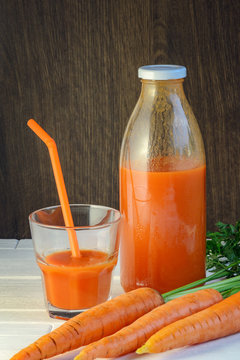 A bottle of carrot juice and a glass of carrot juice with a straw on white wood table against brown wood wall background. Still life of a healthy eating concept