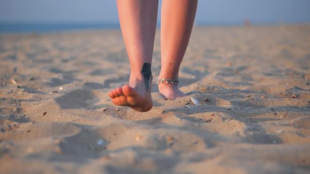 Legs with tattoo of caucasian girl wearing shorts and walking barefoot wet sand sea beach, sun, lens, flare. Slow motion. Woman walks towards to the camera