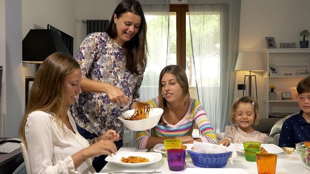 Mother serving spaghetti with tomato sauce to children at the table kissing teenage daughters