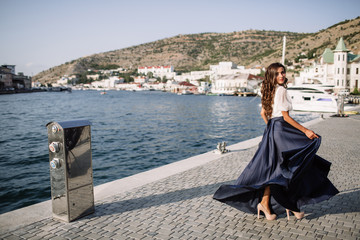 Beautiful girl walking on the pier by the sea near the yacht, boats in the blue long dress