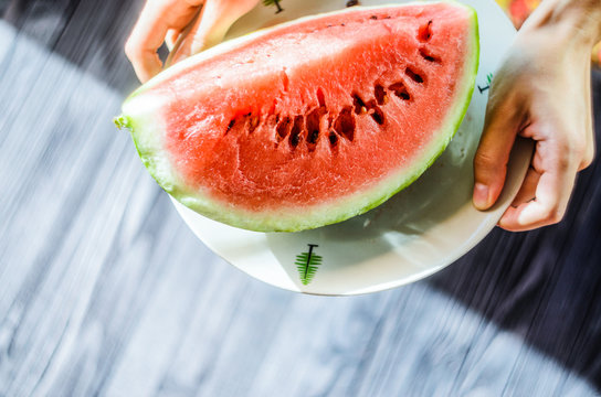 Slice of watermelon on white plate in woman hands ready for eating, top view with blurry black wood table background