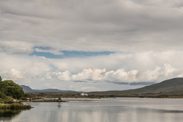 Fototapeta na wymiar Connemara County, Ireland - August 4, 2017: Wide view of Furnace Lake under a gray sky with one blue patch and pronounced low clouds. Green hills and a white house on the horizon.