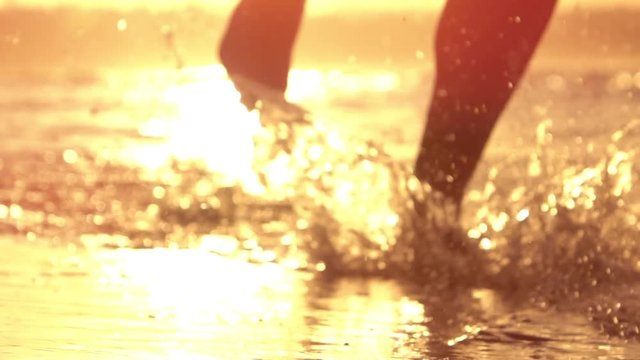 SLOW MOTION CLOSE UP LENS FLARE: Active young man running on sandy beach splashing water at golden sunset. Unrecognizable male runner jogging in shallow ocean water at beautiful yellow morning sunrise