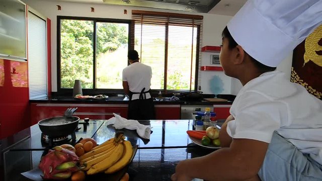 Private Thai chef cooking, his little son in chef's hat nearby sitting on the table in a modern style home kitchen. Making Thai food: mixed vegetables oyster sauce