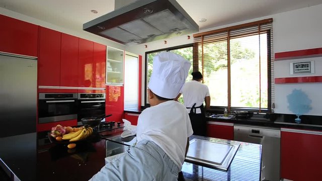 Private Thai chef cooking, his little son in chef's hat nearby sitting on the table in a modern style home kitchen. Making Thai food: mixed vegetables oyster sauce