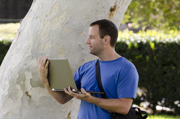 Man looking out while holding a lap top outside in front of a tree trunk.