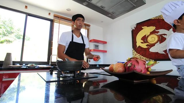 Private Thai chef cooking, his little son in chef's hat nearby sitting on the table in a modern style home kitchen. Making Thai food: mixed vegetables oyster sauce