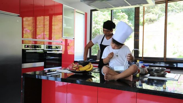 Private Thai chef cooking, his little son in chef's hat nearby sitting on the table in a modern style home kitchen. Making Thai food: mixed vegetables oyster sauce