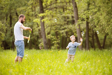 Hipster father and son blowing bubbles outdoors in park.