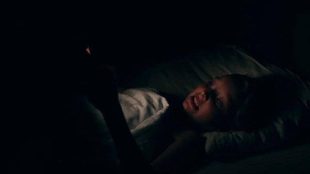 A girl lying in bed watching the horrors on the phone. Night movie viewing. The white girl does not sleep at night. The girl in the bedroom watches the video and is afraid of nightmares.