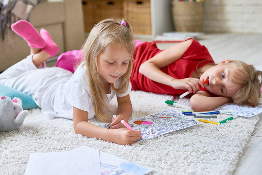 Portrait of two little girls drawing coloring pictures together lying on floor in cozy living room at home