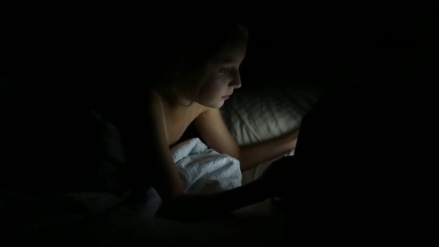 Cute Little Girl Lies in Her Bed at Night, She Holds Tablet Computer. Young, pretty woman using tablet lying on bed at night at home. Young, pretty woman using tablet lying on bed at night.
