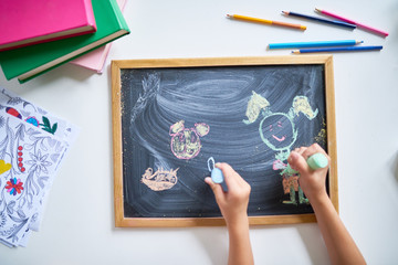 Top view of childrens hands drawing on chalk board with pencils and other supplies on desk around