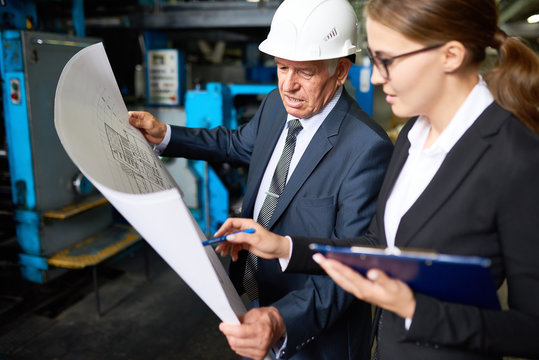 Portrait Of Senior Businessman Looking At Floor Plans Of Modern Factory With Young Assistant Manager In Workshop