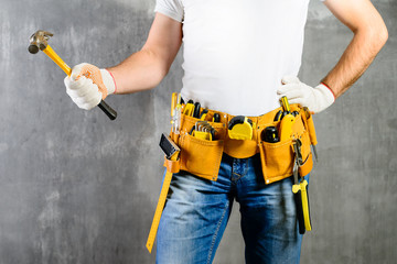 unidentified builder standing in white gloves with a tool belt with construction tools and holding a hammer against grey background. DIY tools and manual work concept.