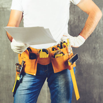unknown handyman with hand on waist and tool belt with construction tools holding the project plane against grey background, toned image. DIY tools and manual work concept