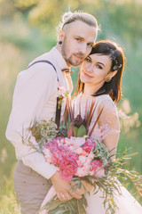 Bride with the hair accessoties and modern-dress groom are holding and hugginh each other in the forest.