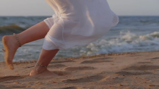 woman's legs with bracelets running on beach in dress at sunset, enjoying freedom during summer holidays vacation travel. Beautiful female girl outside. Slow motion.