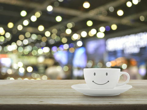 White coffee cup with smile face shape on wooden table over blur light and shadow of shopping mall, Good morning concept