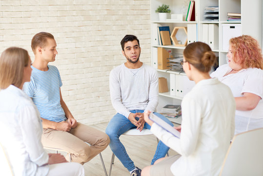 Group of young people sitting in circle sharing mental problems with female psychiatrist holding clipboard