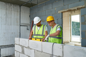 Portrait of two construction workers wearing hardhats and reflective vests  checking concrete block wall using level