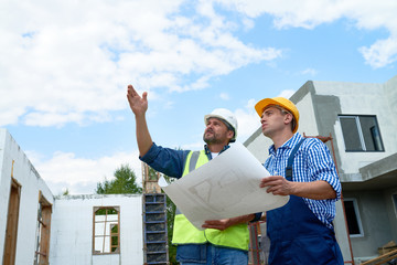 Portrait of two construction workers wearing hardhats discussing floor plans on site, pointing up