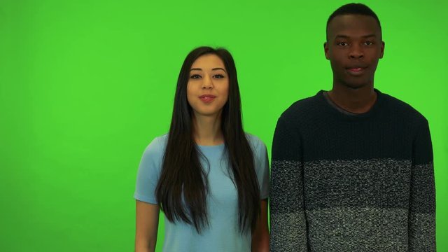 A young black man and a young Asian woman smile and show thumbs up to the camera - green screen studio