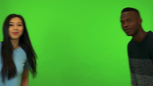 A young Asian woman and a young black man lean into the shot and wave at the camera with a smile - green screen studio