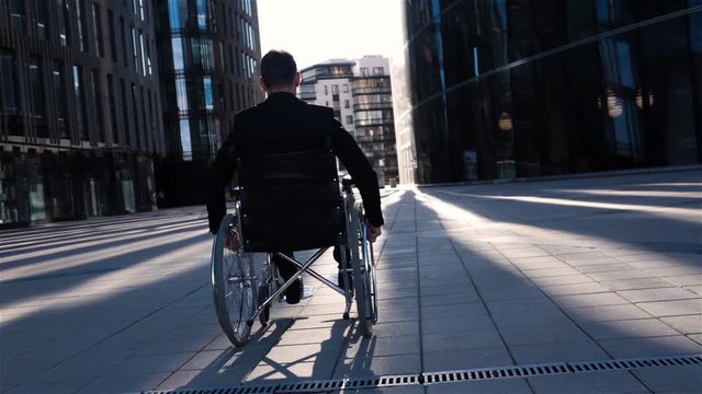 Back view of a invalid cripple businessman in wheelchai with glass building for business deals at bg. He move in his chair quiet in super slow motion. Camera go back with him smoothly. Wide view