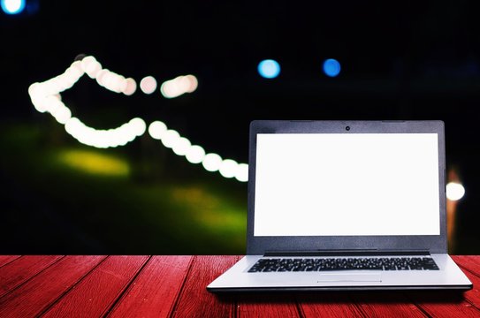 white blank screen laptop computer on wooden desk for presentation with blurred image of night light bokeh of night festival in garden background, copy space, technology concept