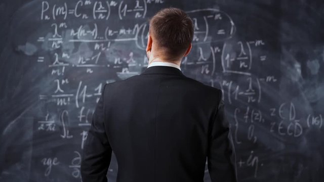 Back middle shot of male against chalk board with mathematics formula equations. He thinking about solutions and try to understand high university task.