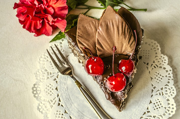 Piece of chocolate cake with cherries and flower China rose on white napkin with a red ribbon on the fork and knife 
