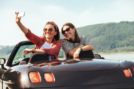 Two female freinds take selfie photo in cabriolet car with beautiful mountain lake view