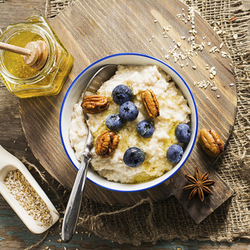 Healthy breakfast: milk porridge from oat bran on skimmed milk with honey, juicy blueberries, pecans on a simple wooden background with a spoon and a jar of honey. Top View.