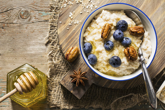 Healthy breakfast: milk porridge from oat bran on skimmed milk with honey, juicy blueberries, pecans on a simple wooden background with a spoon and a jar of honey. Top View.