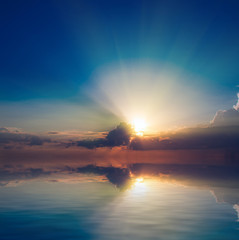 Against the background of a blue sky, a sunset with sunbeams and a sea without waves.