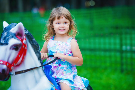 Cute little girl having fun outdoors in the summer park and riding a toy horse