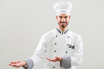 Smiling chef showing welcome gesture on gray background.