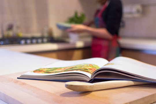 Cook book put on a kitchen table with a wooden spoon in the foreground. A  young woman with apron is cooking in the Kitchen. Stock Photo | Adobe Stock