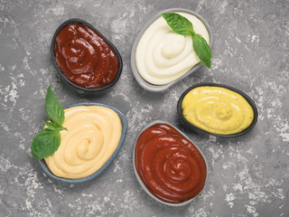 Top view of classic sauces set in trendy plates on gray concrete background. Sauces set - salsa, mustard, ketchup, mayonnaise, cheese sauce, and basil for serve. Flat lay.