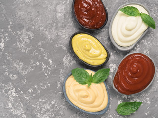 Top view of classic sauces set in trendy plates on gray concrete background. Sauces set - salsa, mustard, ketchup, mayonnaise, cheese sauce, and basil for serve. Flat lay. Copy space.