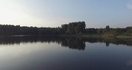 Fototapeta premium Camera flight over the lake, clear like a mirror. It reflects the trees on the shore, green grass, wooden house and blue sky.