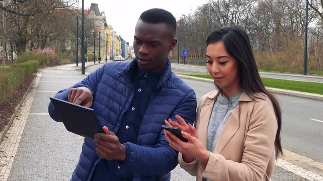 A young black man and a young Asian woman work on a tablet and a smartphone in a street in an urban area, he shows her his screen while explaining