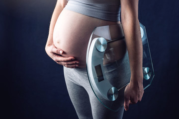 Pregnant woman holding scales and weigh keeping one hand on a belly. Concept of weight loss and control during pregnancy